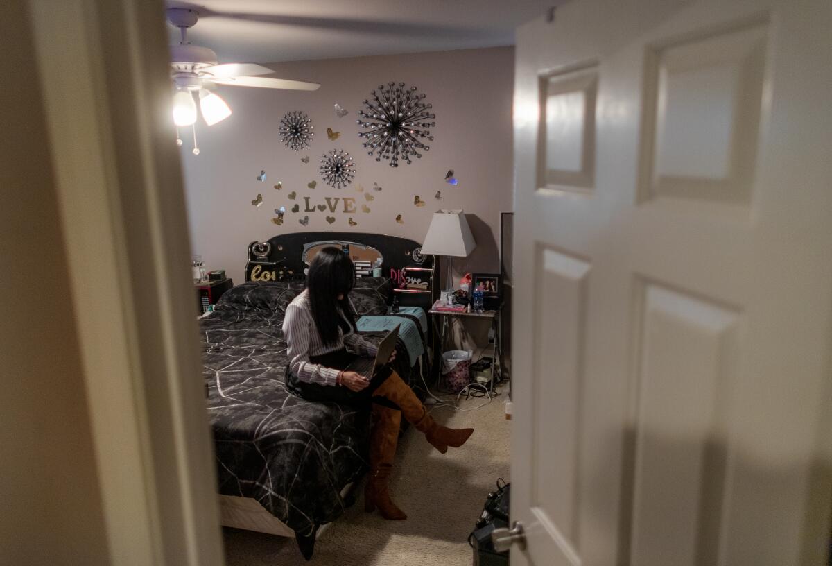 A view from the door of a woman with long black hair covering her face sits on the side a bed and looks at a laptop. 