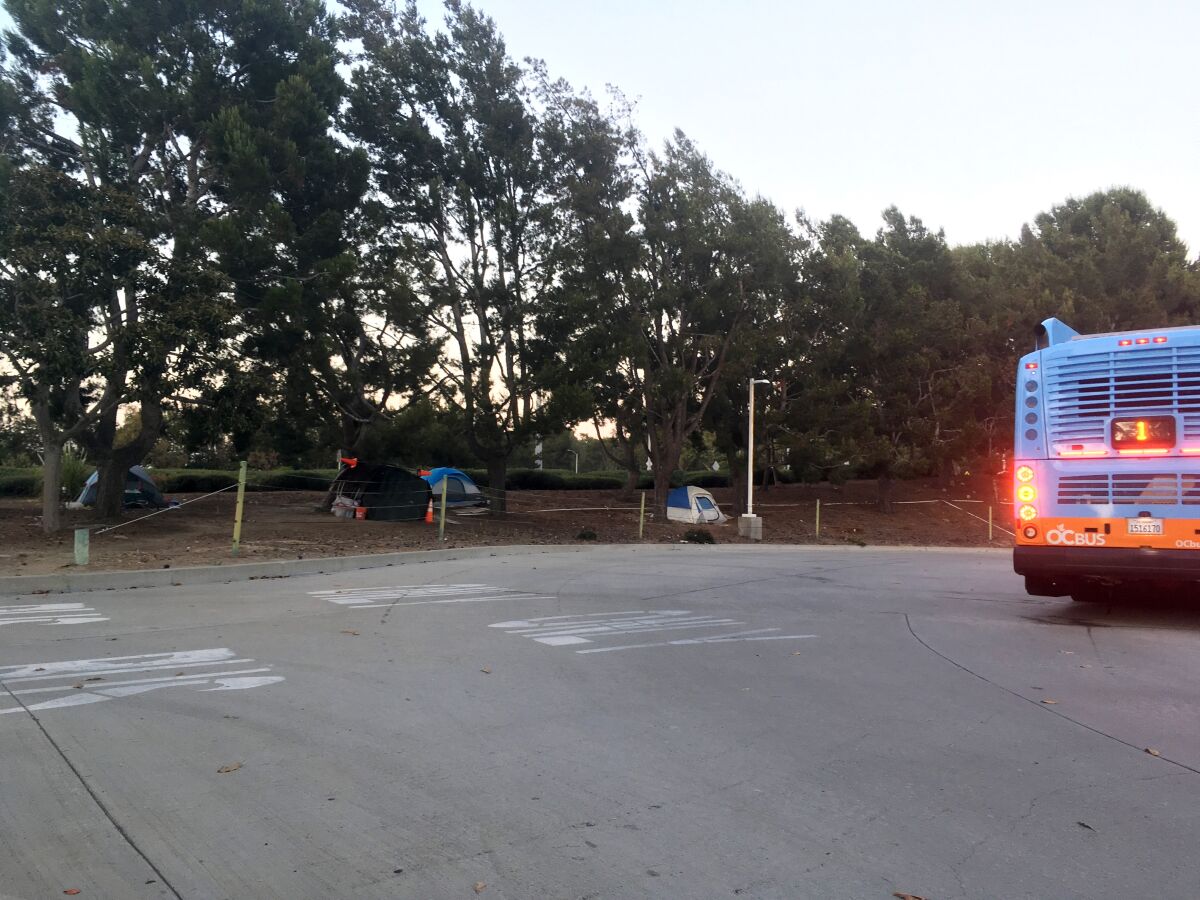 Tents sheltering homeless people sit on a tree-shaded slope on the north side of the Newport Transportation Center at San Joaquin Hills Road and MacArthur Boulevard.
