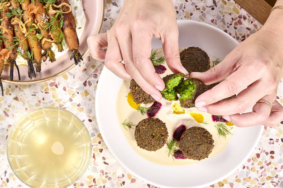 An overhead photo of hands ripping open fresh, green falafel next to a plate of roasted carrots and a glass of wine.