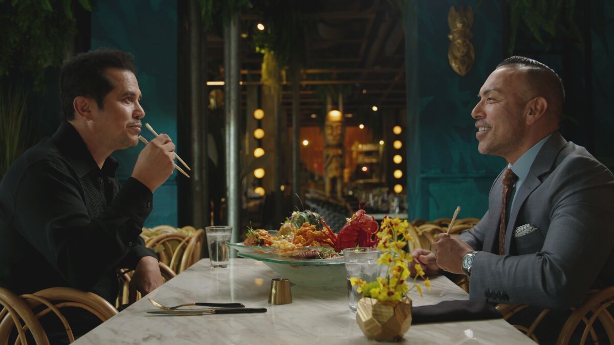 John Leguizamo and Juan Chipoco sit at a table eating and talking