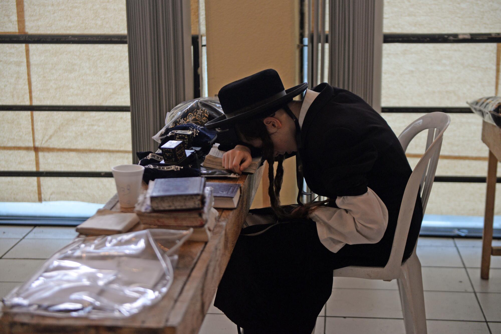 A person with earlocks and dark robe and hat leans down on a book atop a table 