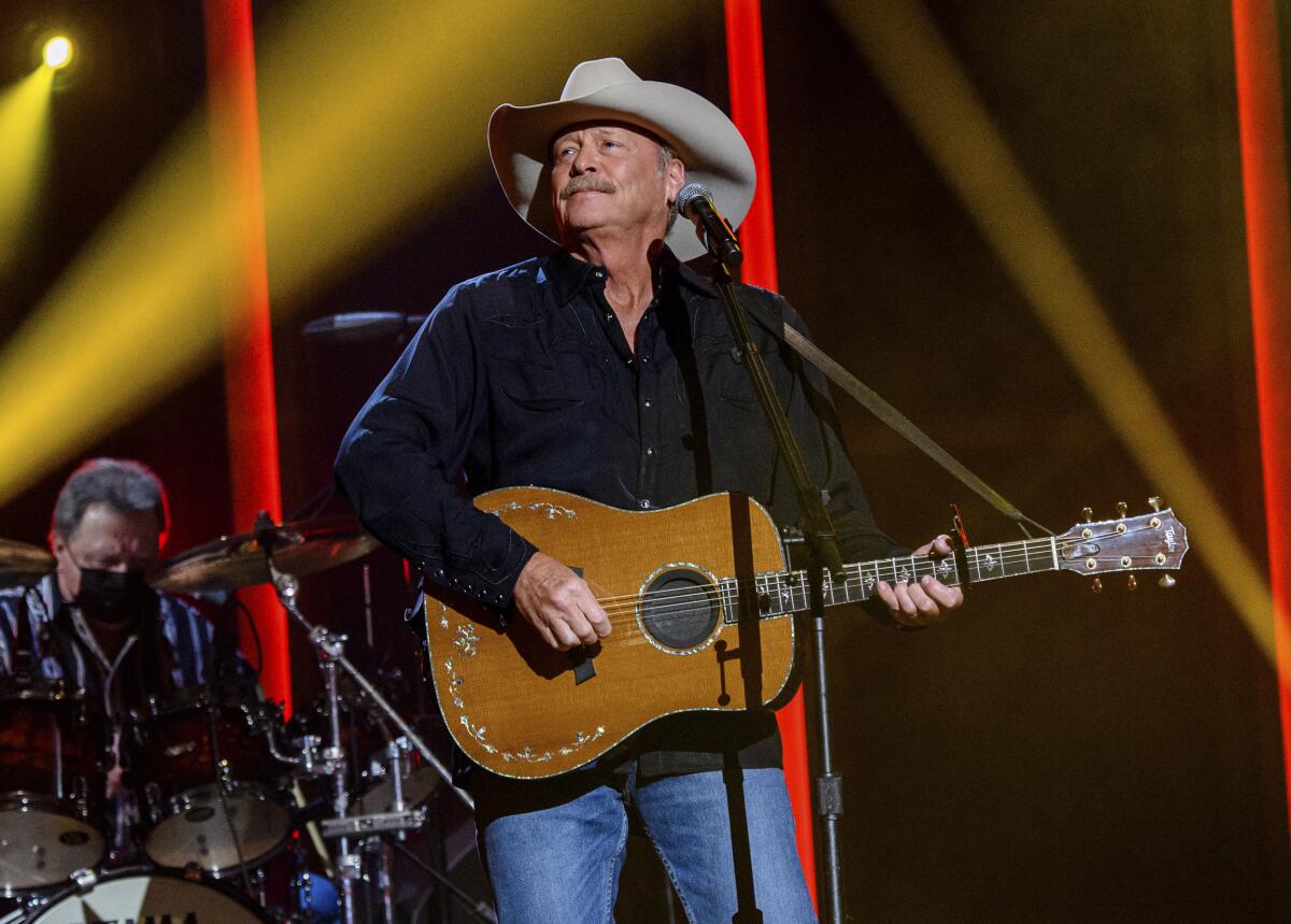 A man wearing a black long-sleeve button down and blue jeans with a cowboy hat plays guitar on a stage.