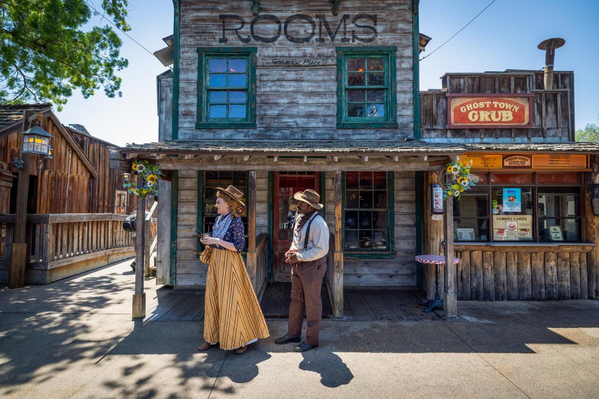 A woman in a long skirt and a man in suspenders stand in front of an Old West backdrop.