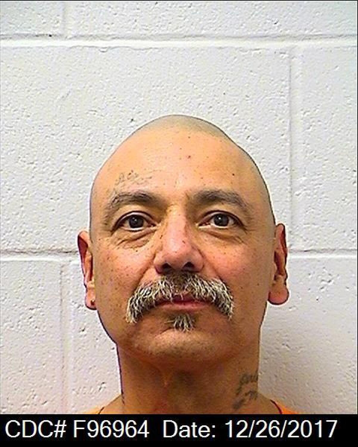 A mugshot of a man with a shaved head and salt-and-pepper mustache photographed against a white block wall.