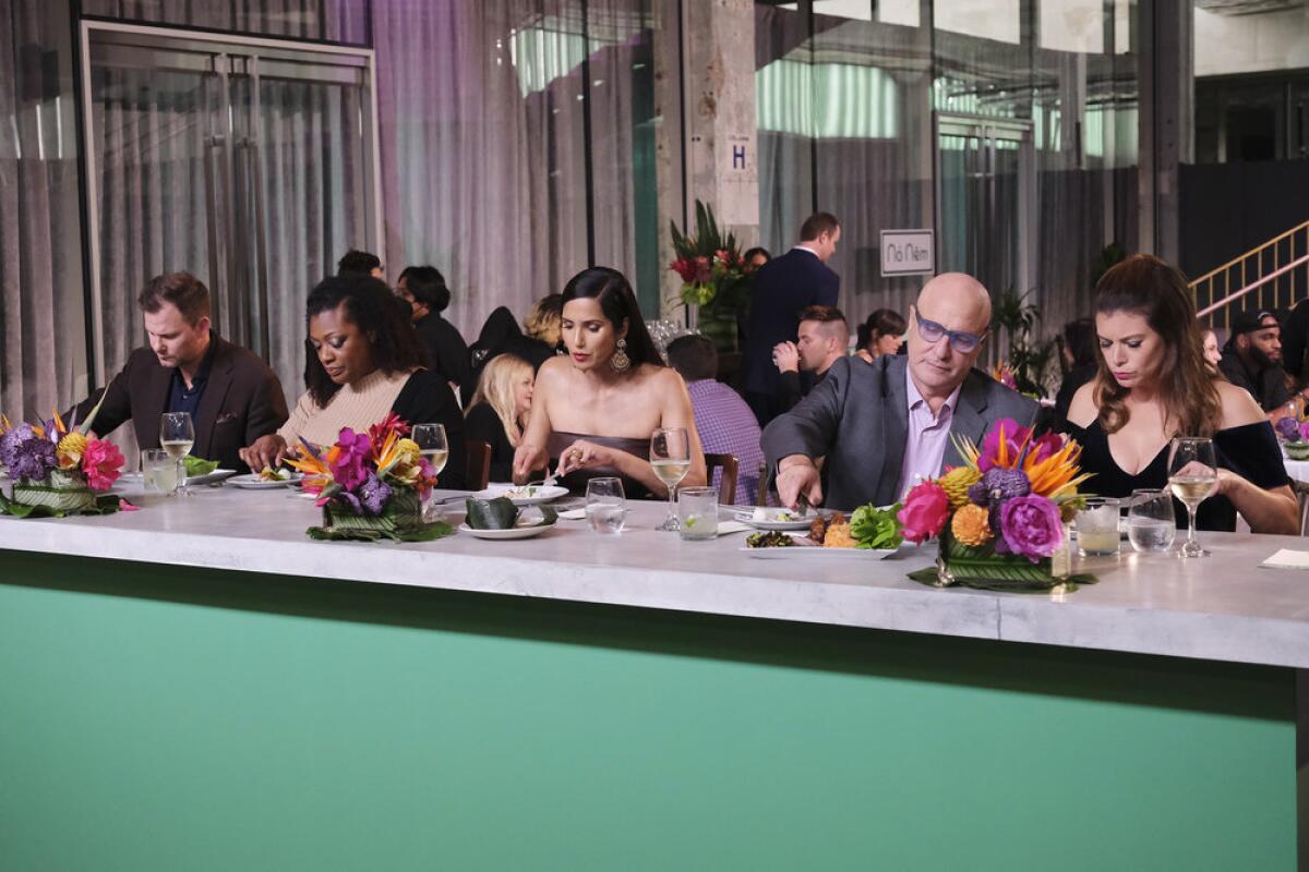 Three women and two men sample a dish at a restaurant table with flower arrangements on it.