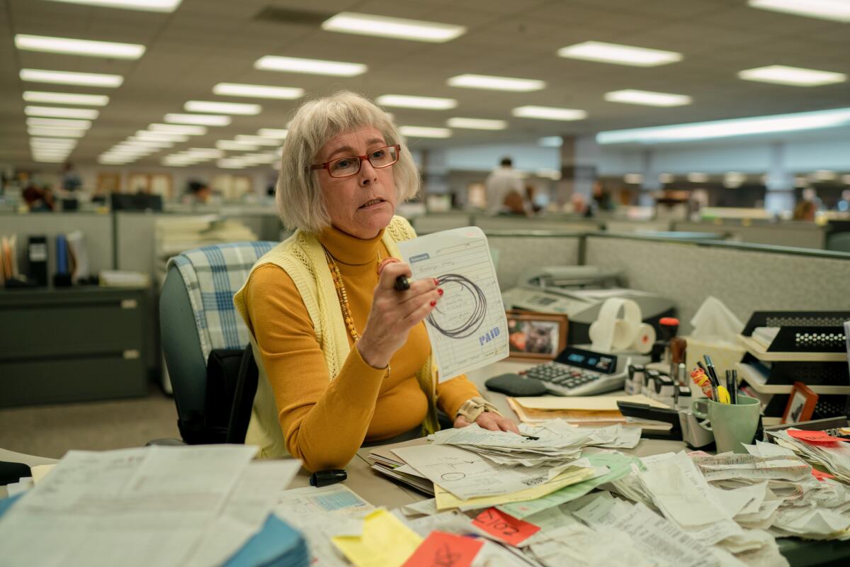 A frumpy woman sits at a desk piled with paperwork.