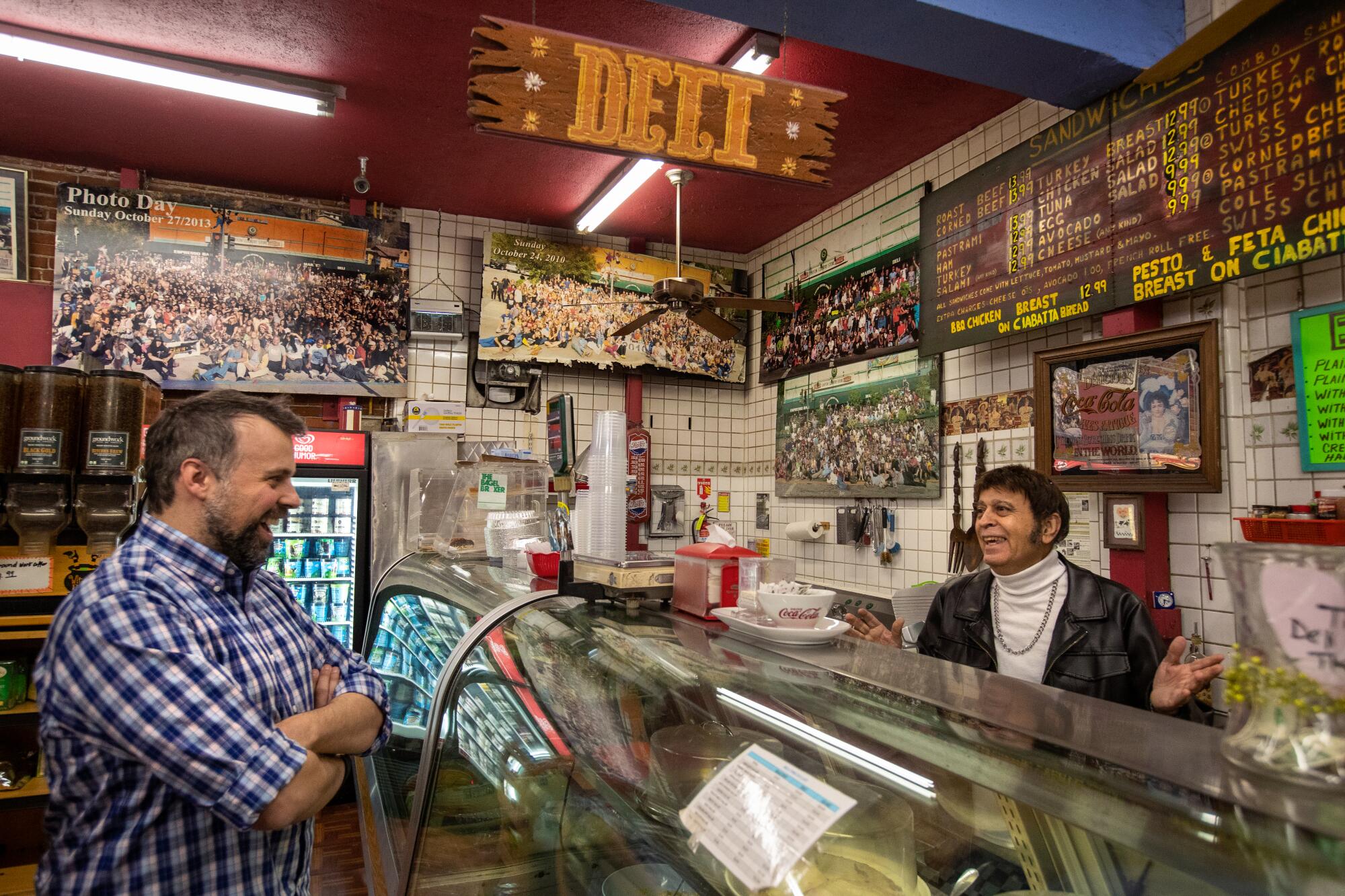 Two men laughing stand on either side of a deli counter.