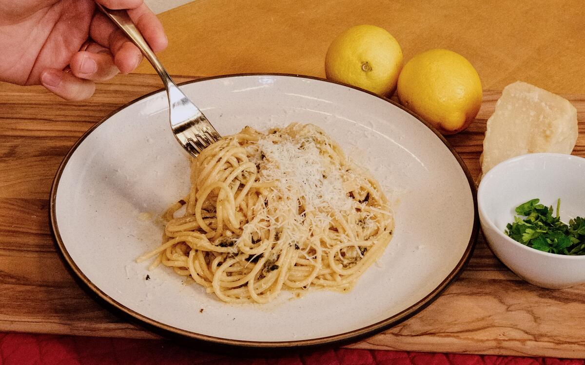 A hand lifts a forkful of spaghetti al limone from a plate in the L.A. Times Test Kitchen. Lemons, herbs and cheese behind.