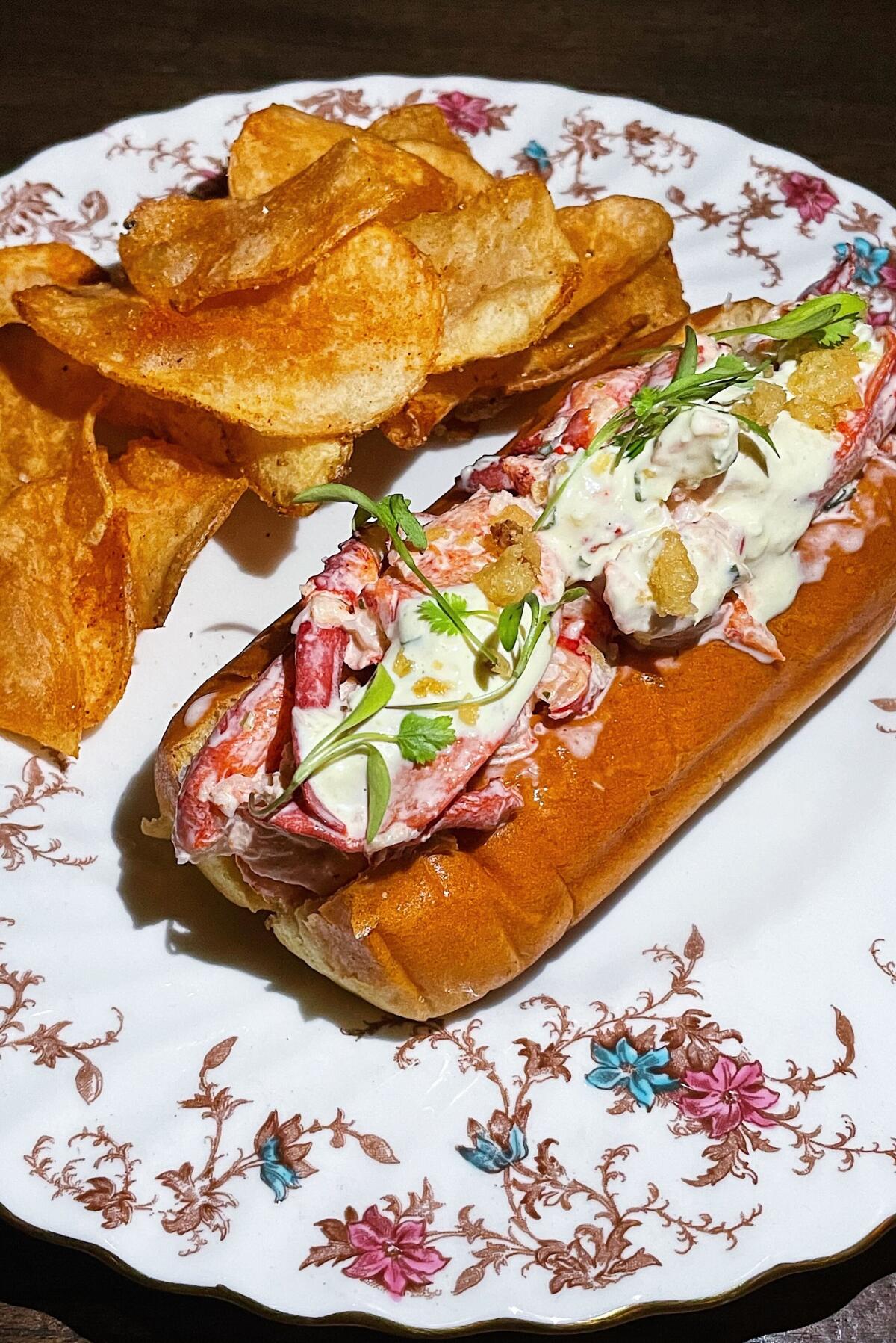 A lobster roll topped with tempura leeks on a flowered plate, with a side of chips