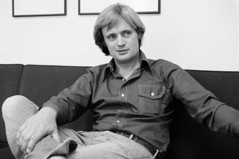 A black-and-white image of David McCallum lounging in a couch in a denim outfit