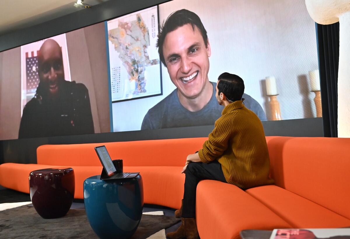 Two men on a big screen talk with a man on a couch in a room.