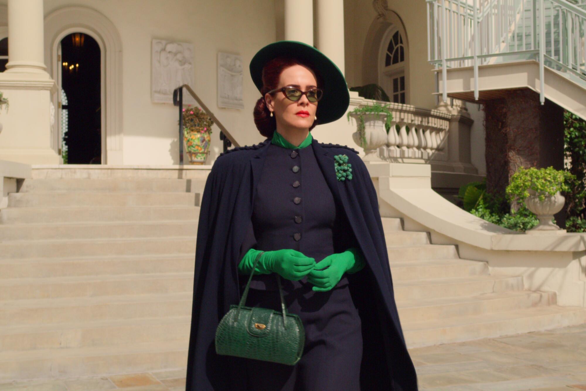 A woman in a black cape and hat, sunglasses and green gloves outside a building on a sunny day.