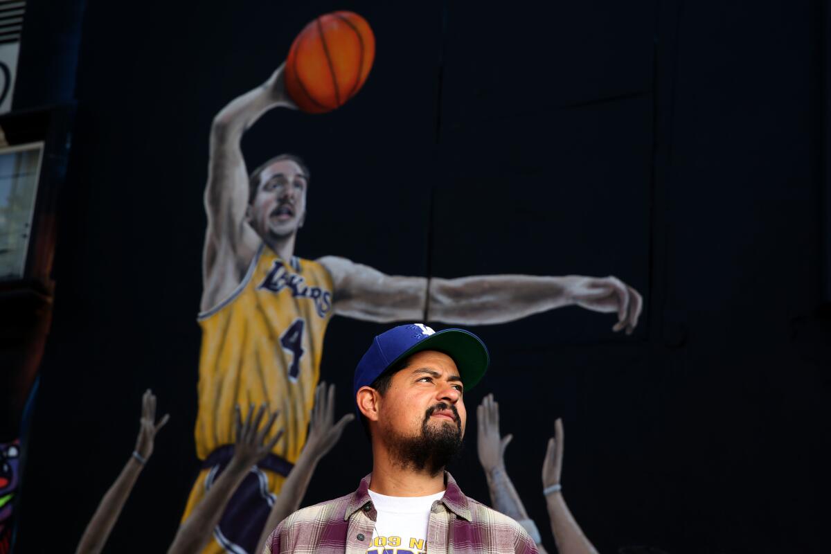 Artist Gustavo Zermeo Jr. in front of a mural he painted of Lakers guard Alex Caruso and other basketball players of the West Coast in the parking lot of Sportie LA.