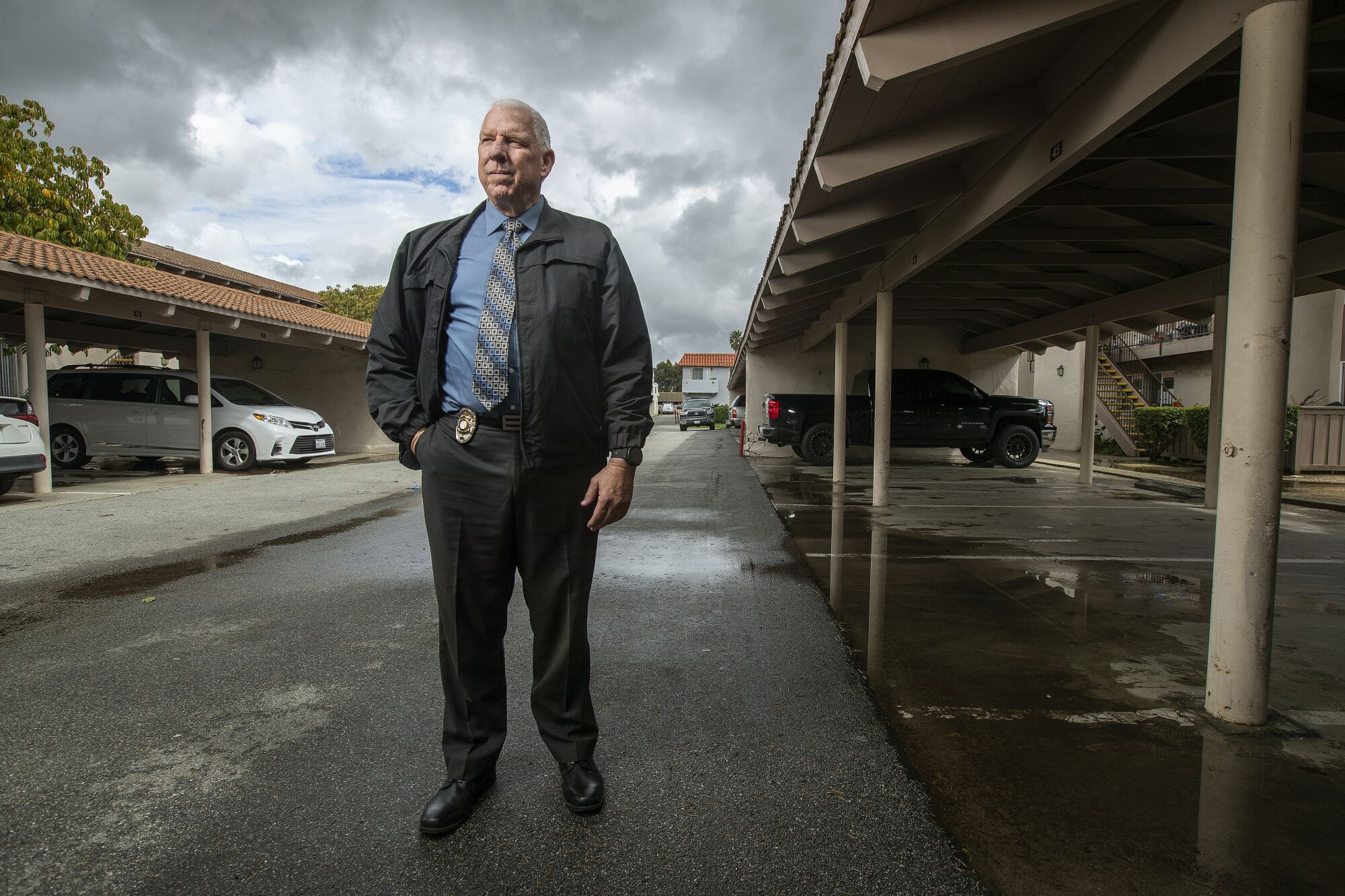 A man wearing a dark jacket and detective's badge on his belt stands beneath a gray sky.