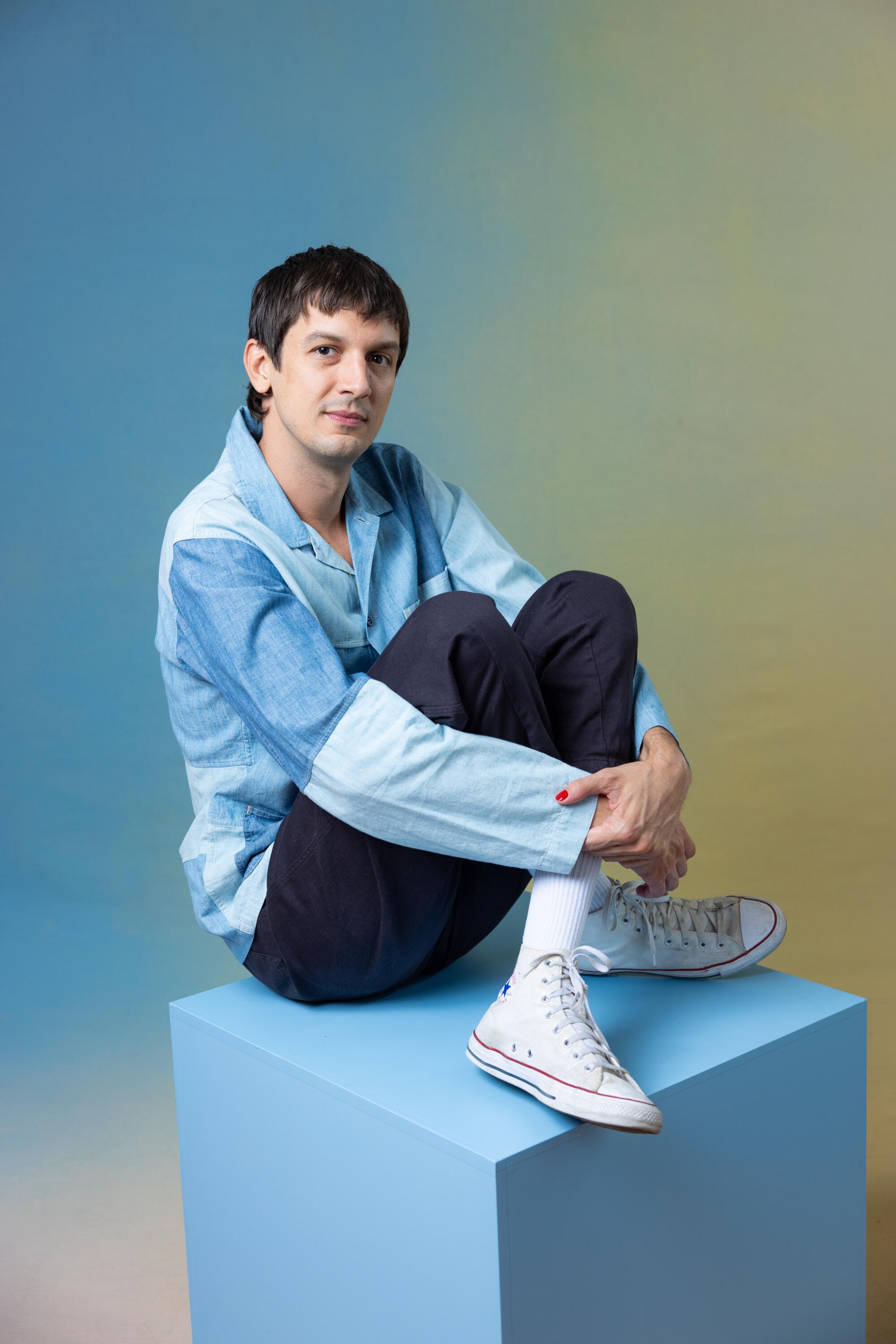 Josh Sharp wears a blue shirt and dark pants while sitting on a blue cube.