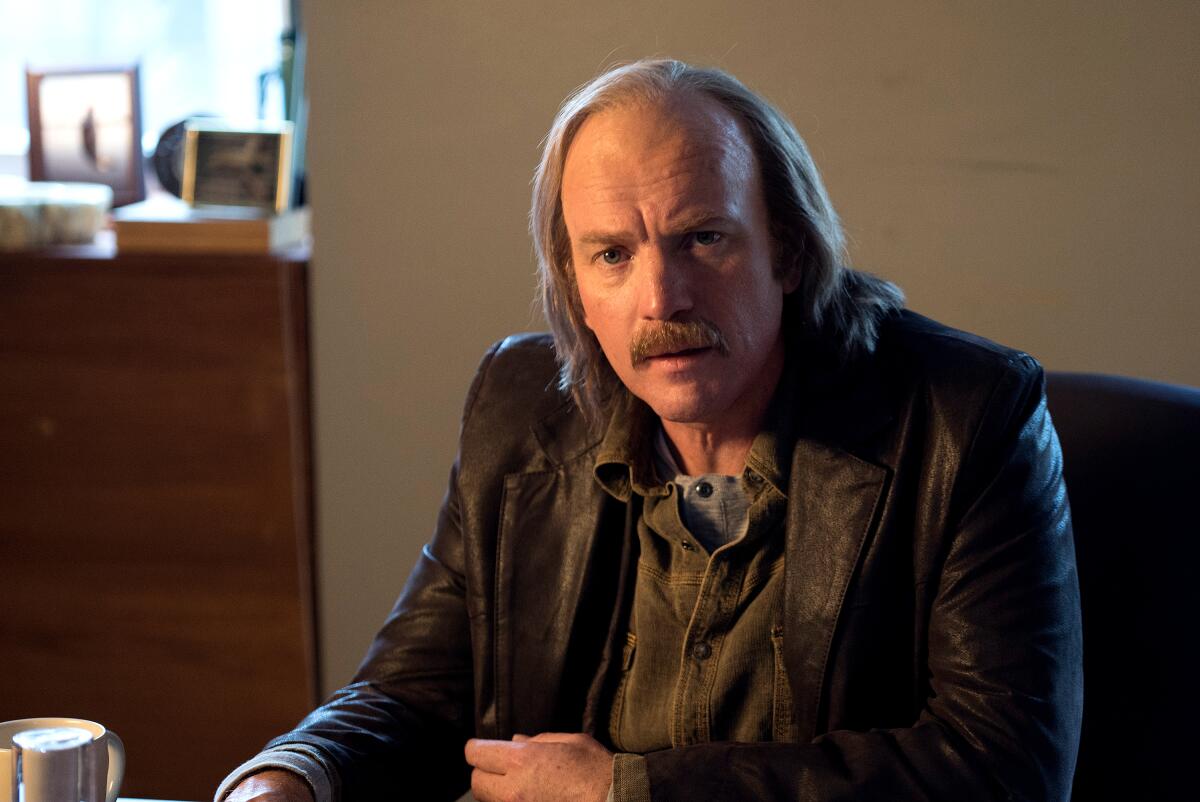 A balding man in a leather jacket sits in an office in "Fargo," Season 3.