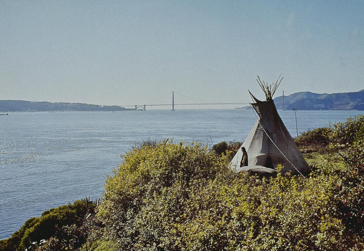 A color photo shows a man standing before a tepee on the Alcatraz shore with the Golden Gate Bridge in the distance