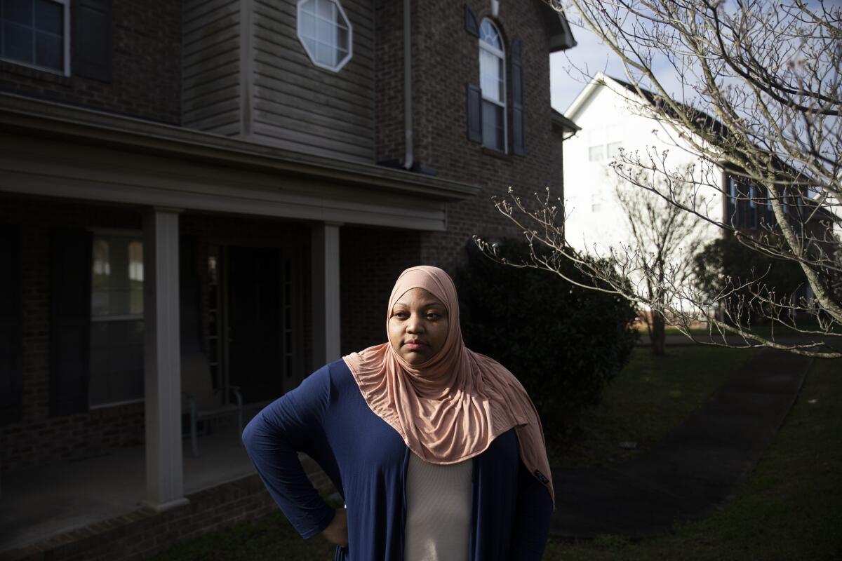 A woman in a hijab standing outside a two-story home