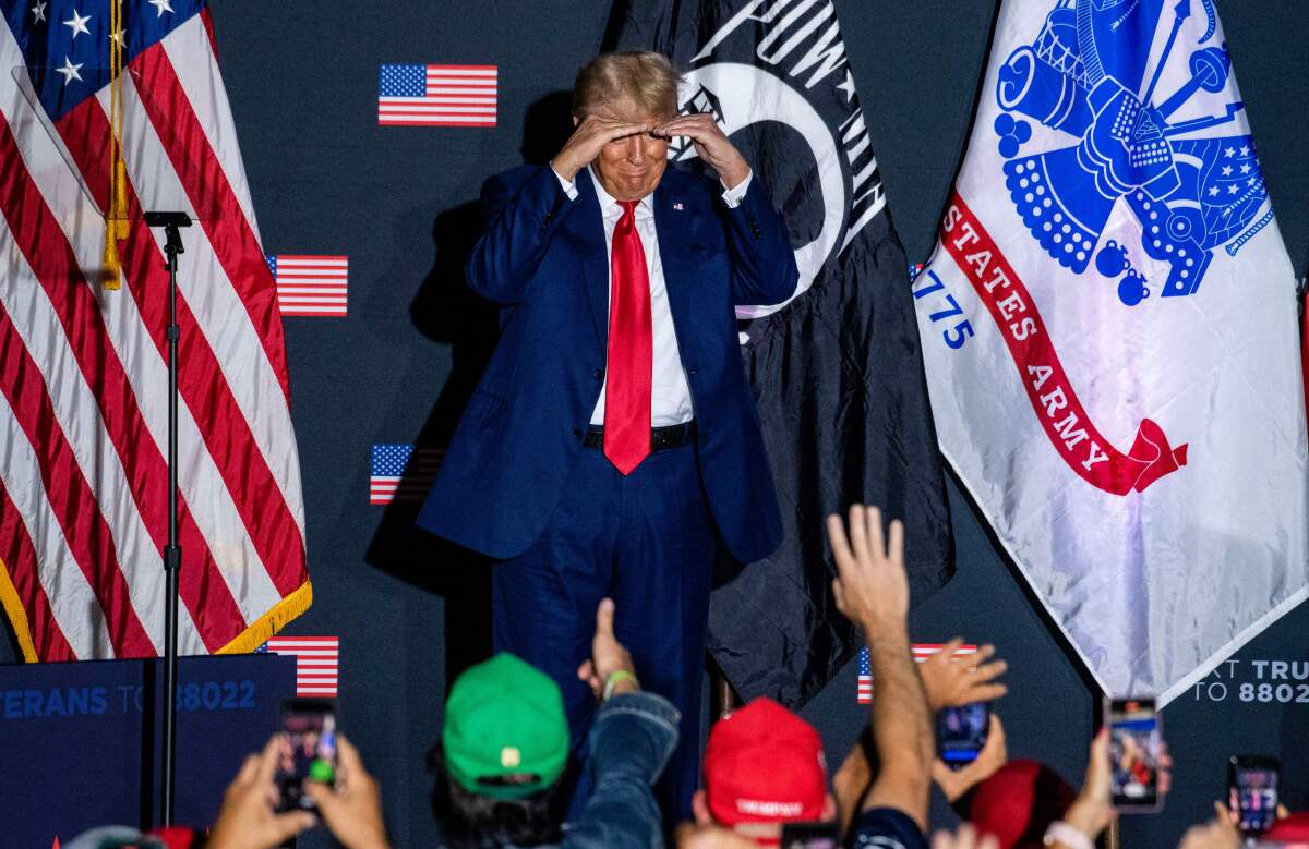 Ex-President Trump, hands shielding his eyes, looks into a crowd from a stage decorated with American, POW-MIA and Army flags