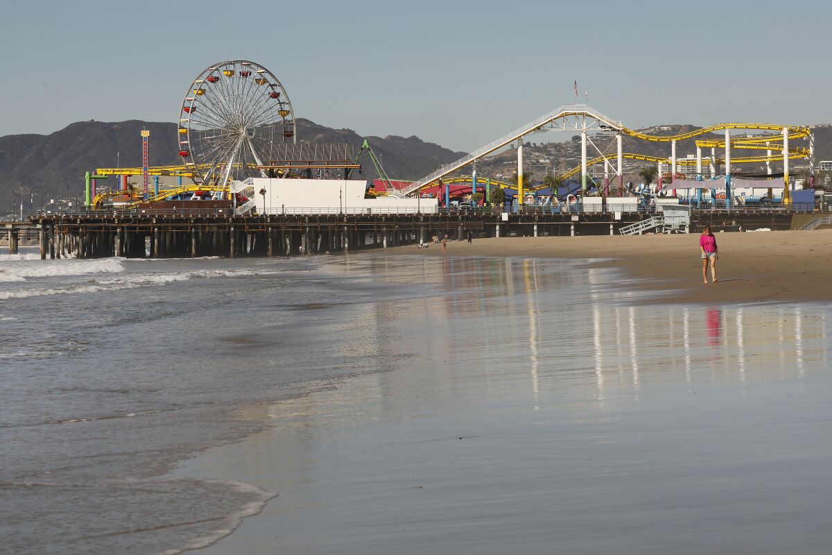 A person walks along the beach with the Santa Monica Pier in the background.