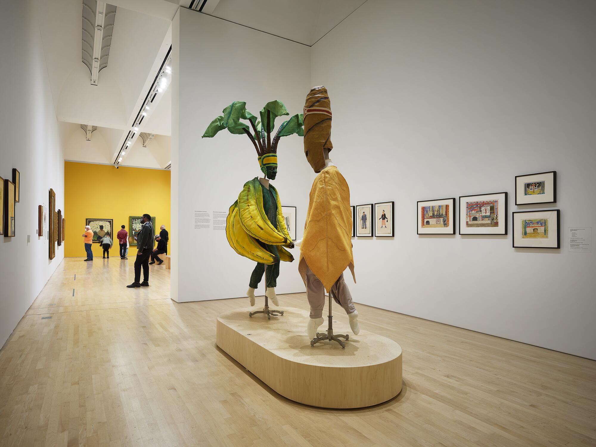 A gallery view shows a banana costume and a cigar costume on a plinth before a wall of drawings by Diego Rivera