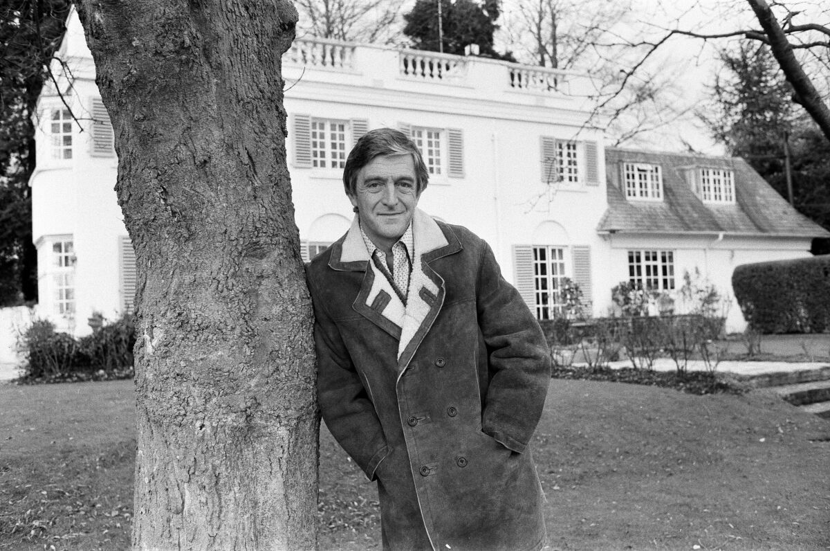 A man leans against a tree in front of a white house.
