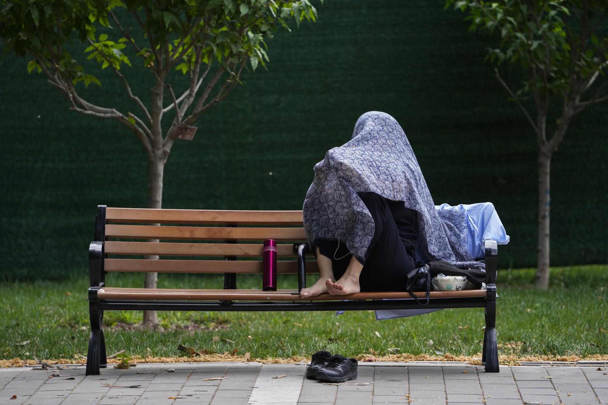 A barefoot woman, using a scarf to shield from the sun, sits on a bench.
