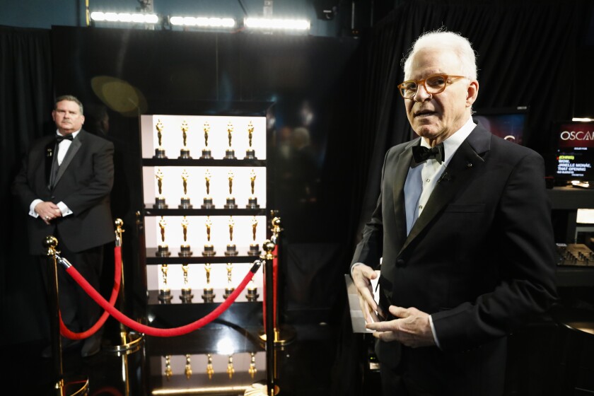 A man in a tuxedo stands by a velvet rope in front of Oscar statuettes in a case.