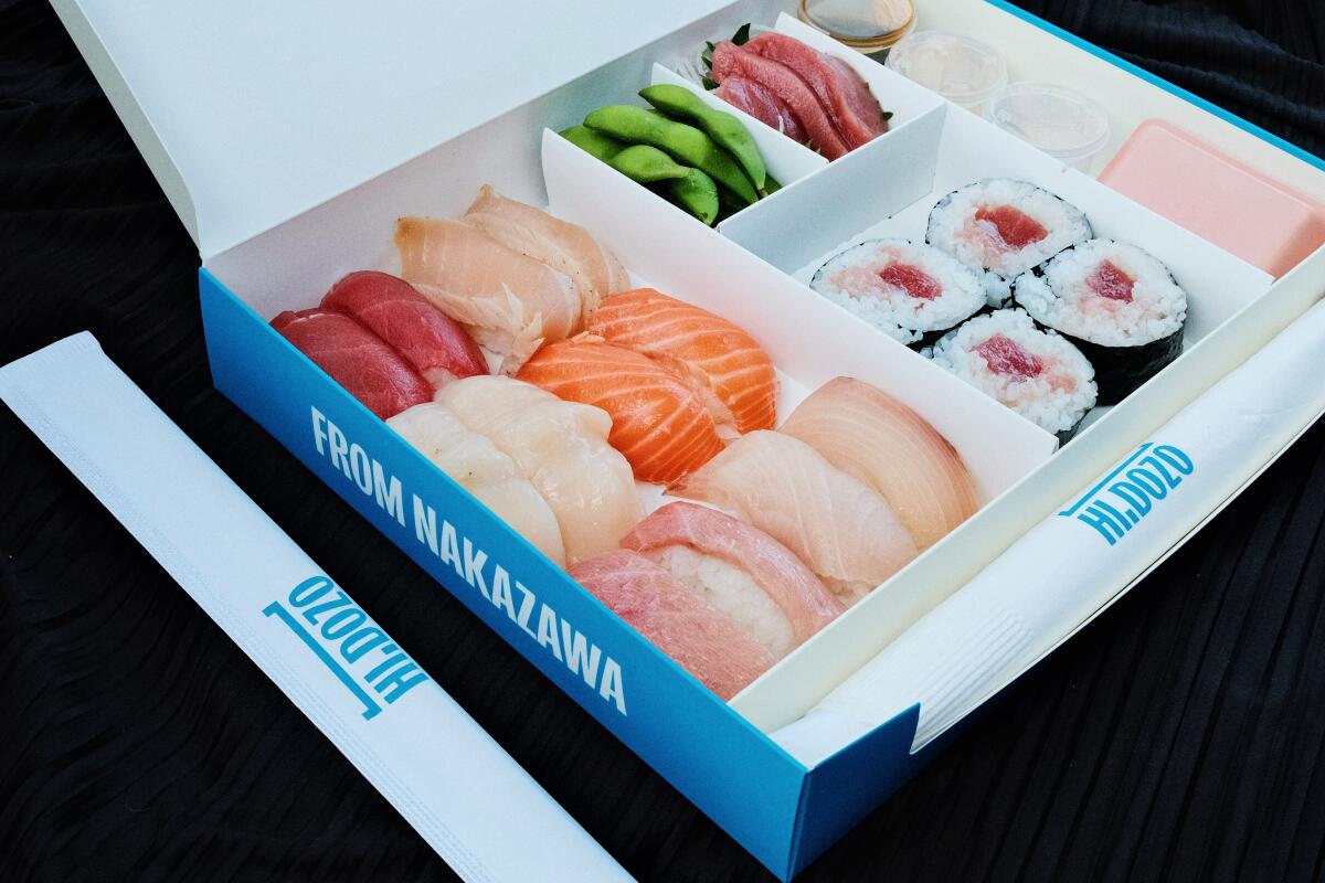 An angled photo of a white and blue paper box of sushi featuring pieces of nigiri, maki, sashimi and edamame.