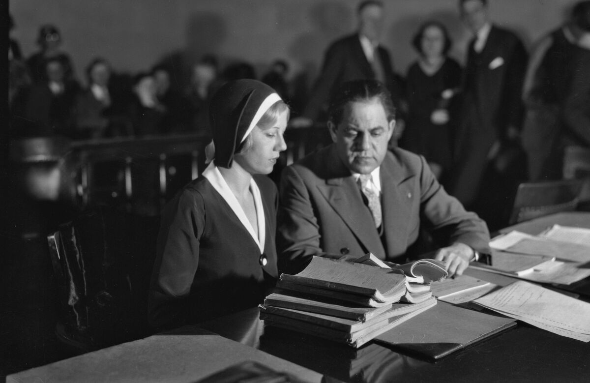 A woman in a dark dress and matching hat sits beside a man in a suit. The table in front of them is stacked with papers.