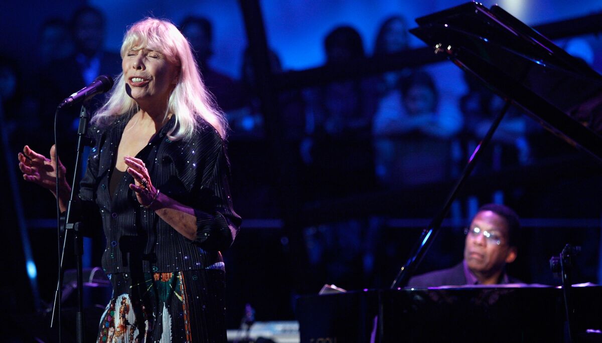Joni Mitchell singing into a microphone onstage. Behind her, Herbie Hancock plays the piano.