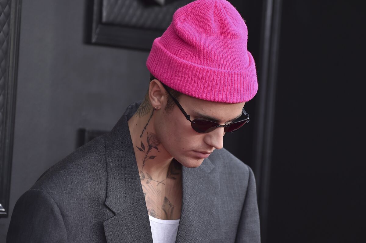 A man in gray suitcoat with neck tattoos, dark glasses and a pink knit cap looks downward