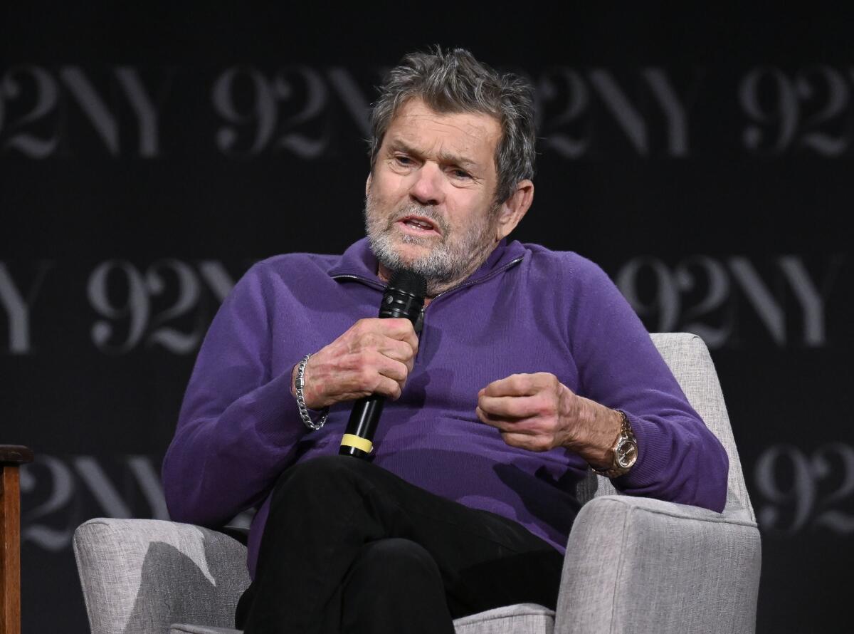 Jann Wenner sits in an armchair holding a microphone.