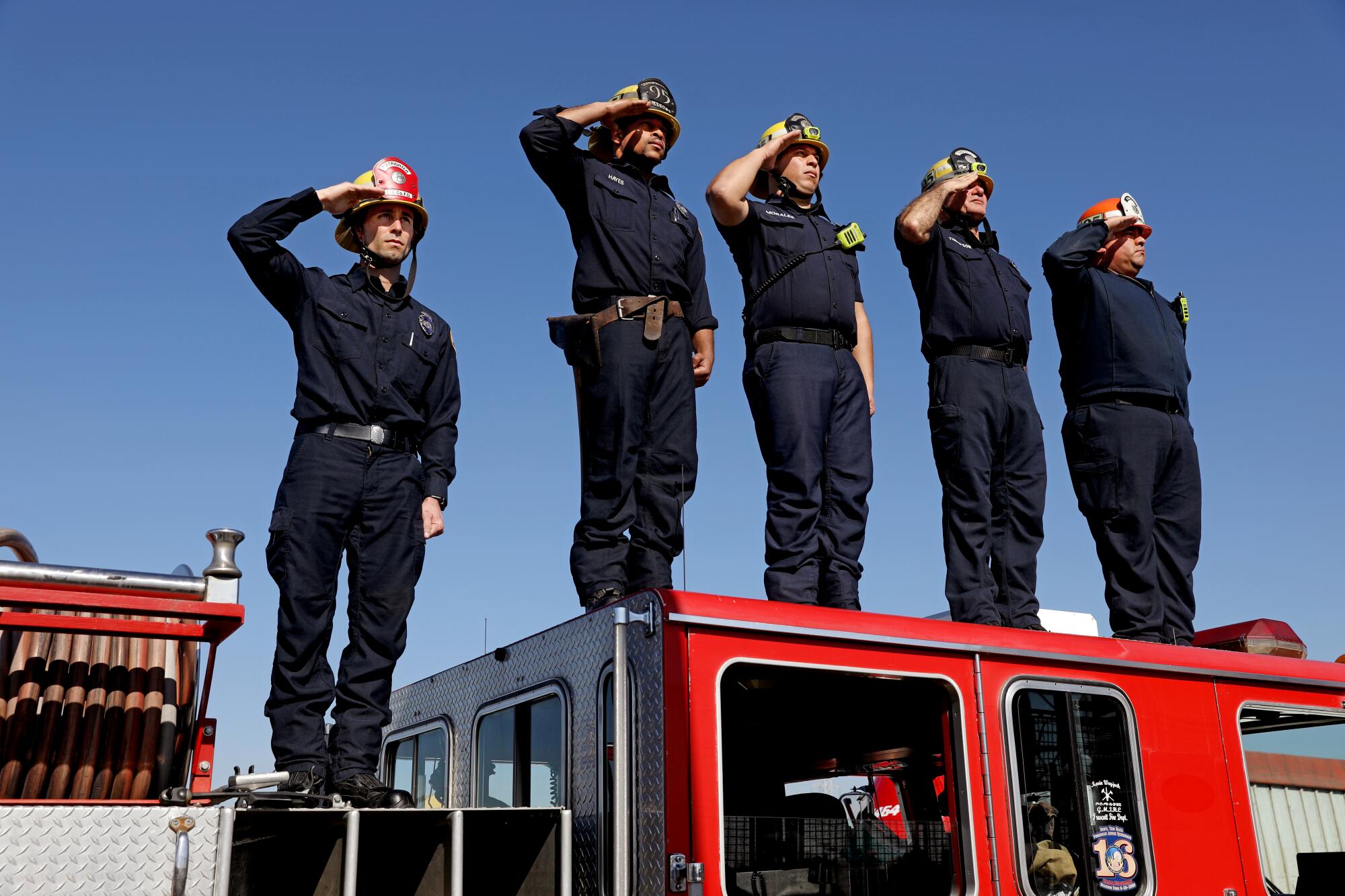 Five firefighters stand atop a firetruck while saluting