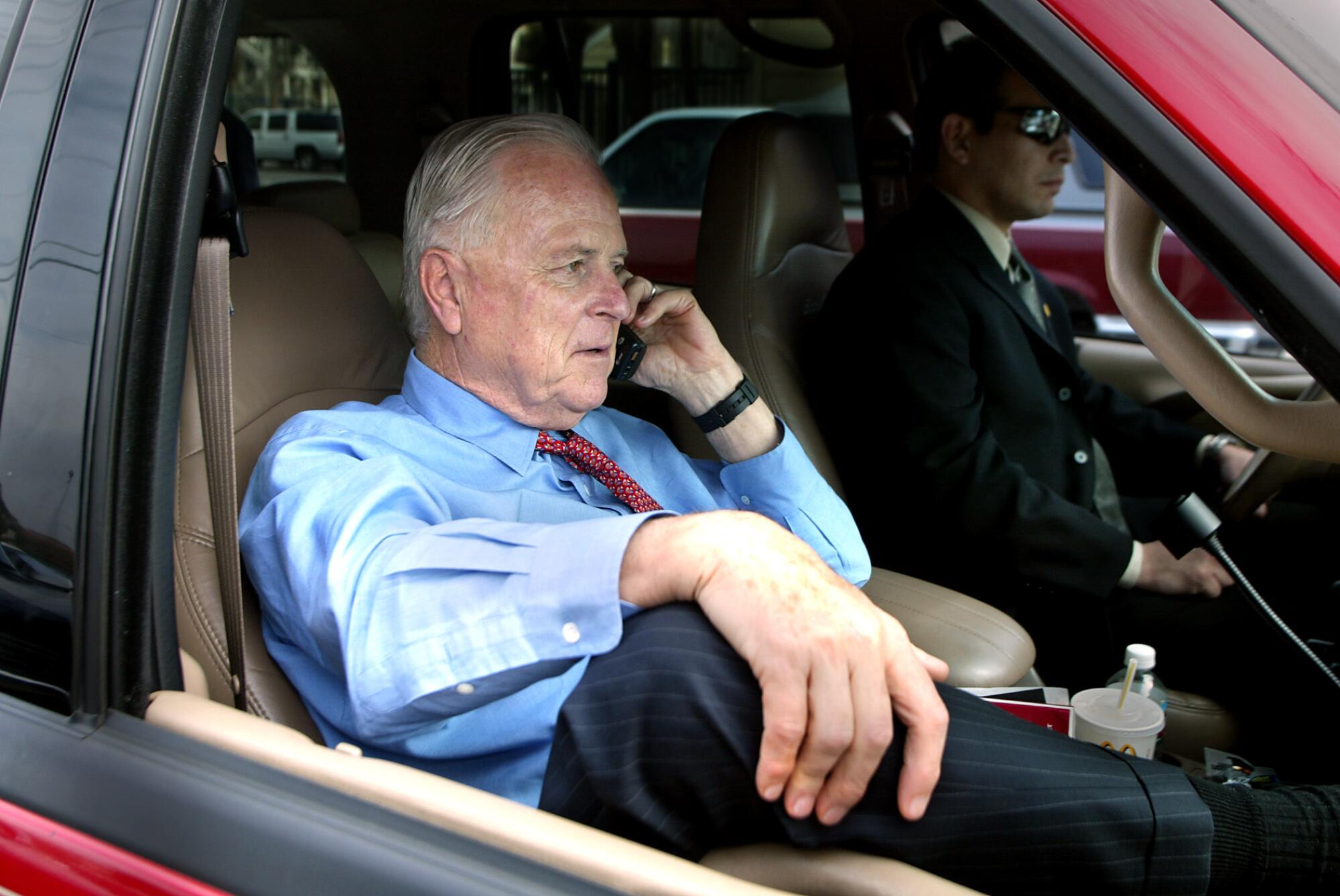 Richard Riordan talks on a cellphone sitting in the passenger seat of a car
