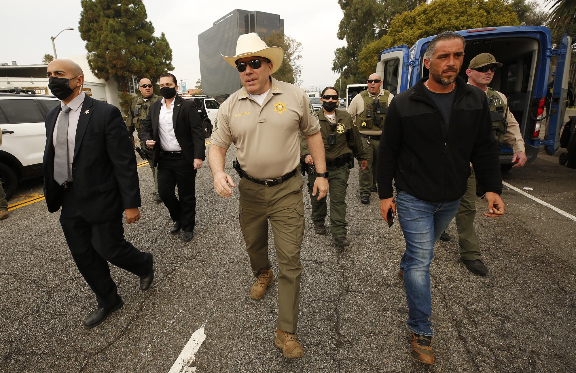 Alex Villanueva, wearing a cowboy hat, walks with others while touring the Veterans Row homeless encampment