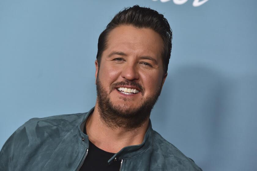 Luke Bryan smiles against a light blue background while wearing a light blue moto jacket