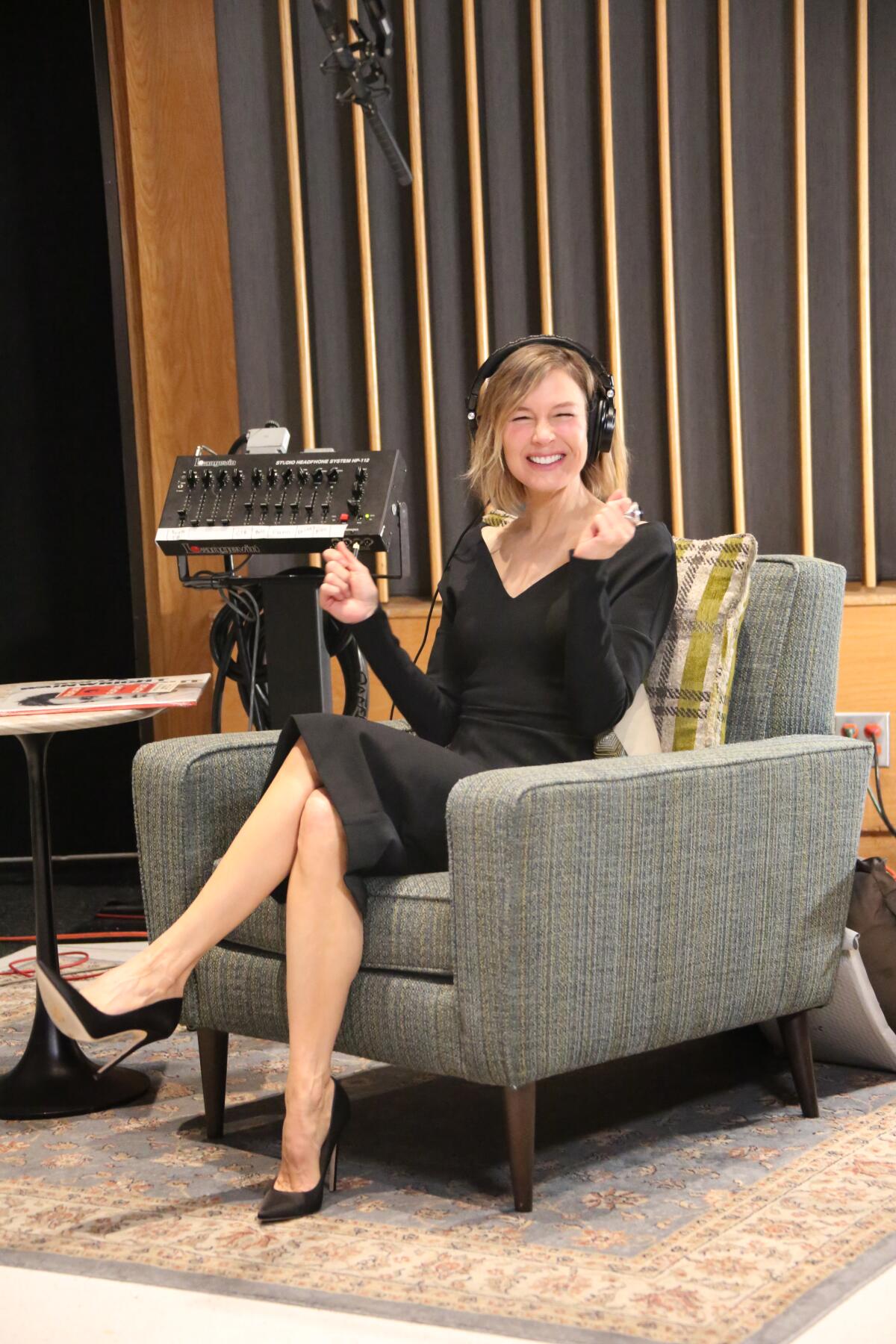 Wearing headphones, Renee Zellweger sits in an armchair with her legs crossed at the knees.