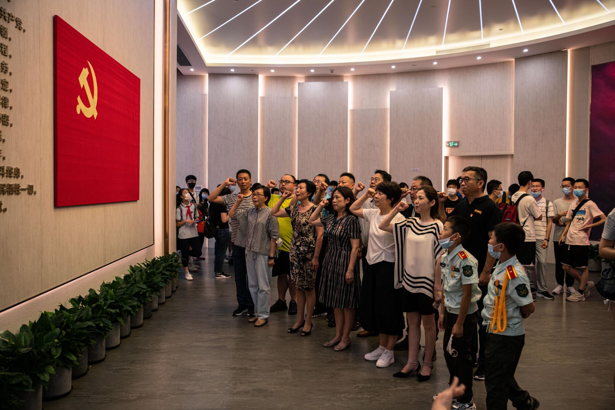 A group of people raise fisted hands as they stand before a red flag with a yellow hammer and sickle symbol on a wall 