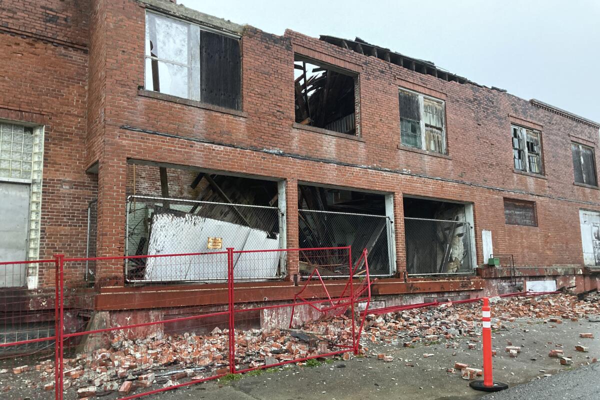 A red-brick building with blown-out glass windows, a lot of brick fallen around, and a partially caved-in roof.