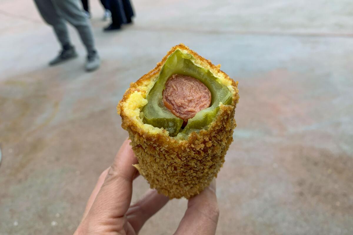 A hand holds an open corn dog, revealing the pickle-wrapped hot dog inside the bun
