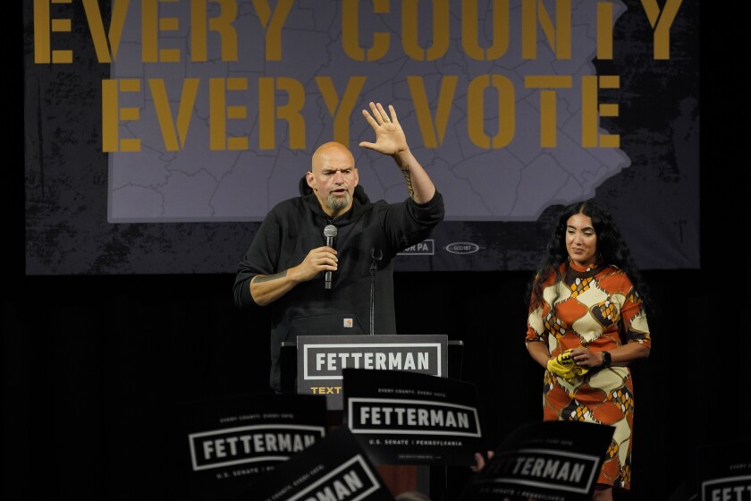 John Fetterman, wearing a hoodie, waves and holds a microphone on a rally stage while his wife stands next to him.