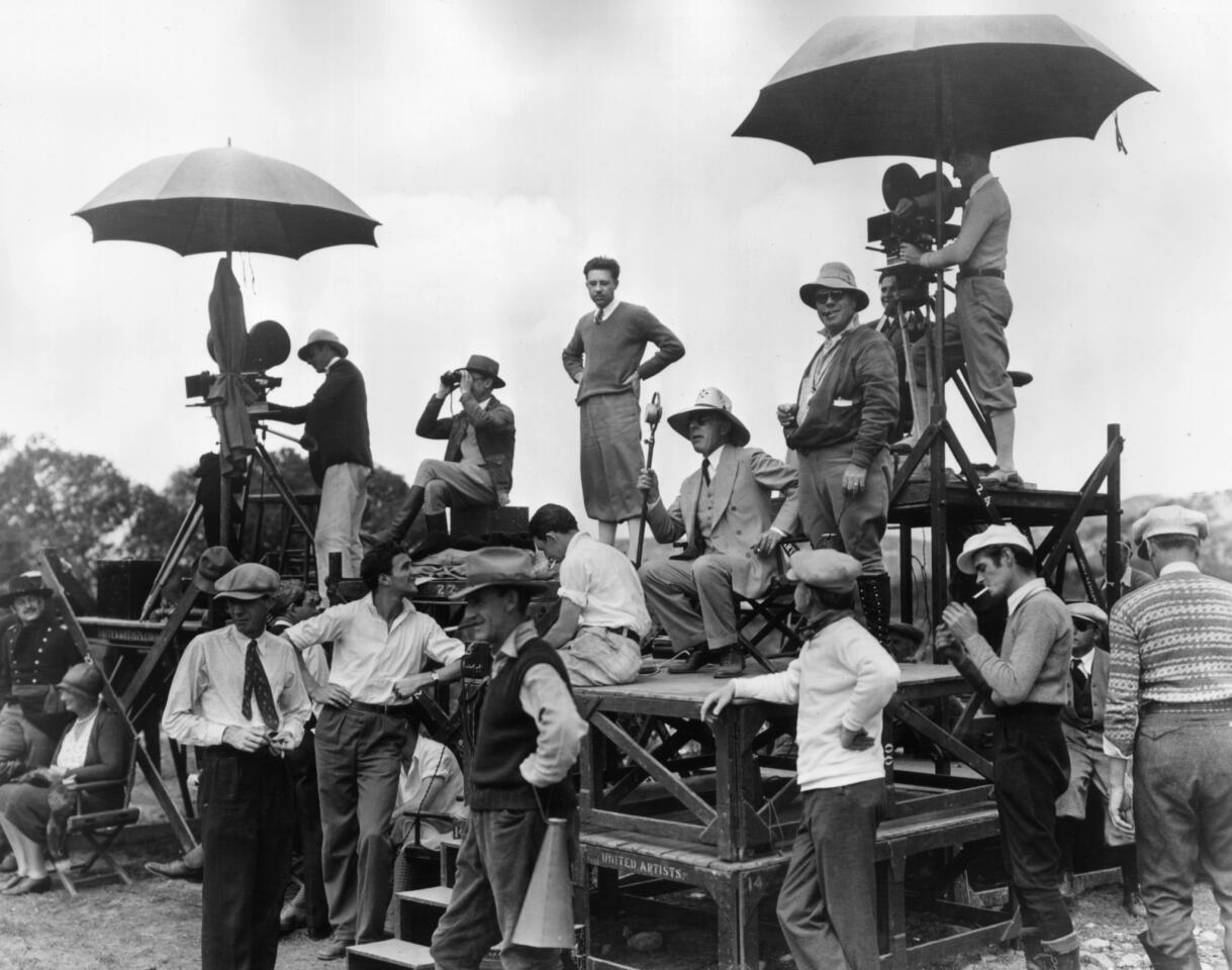 Circa 1915: American director David Wark Griffith, center in suit and tie, sits on a platform surrounded by crew members on the set of a film.