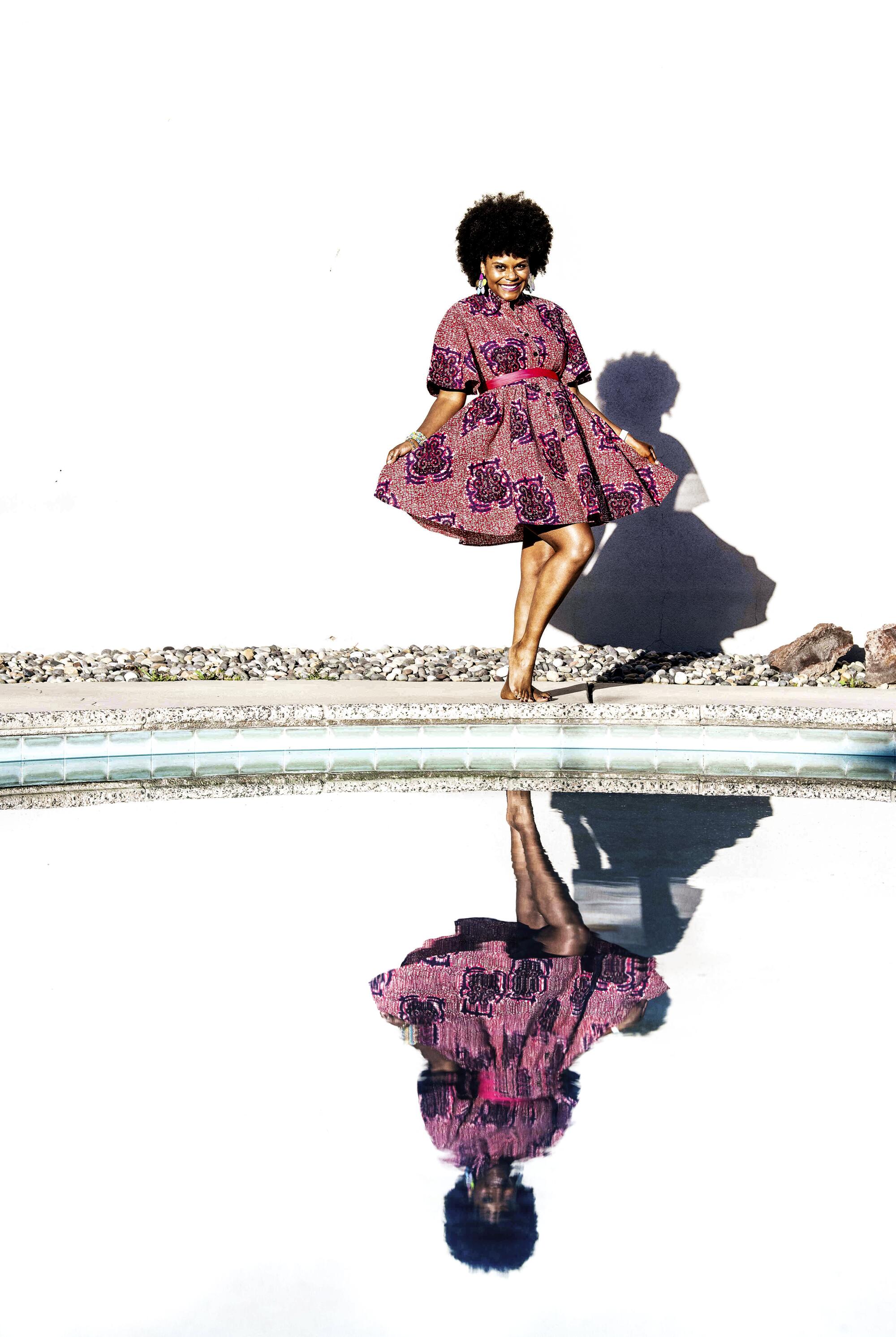 Tabitha Brown poses over a reflection of herself in a body of water outdoors.