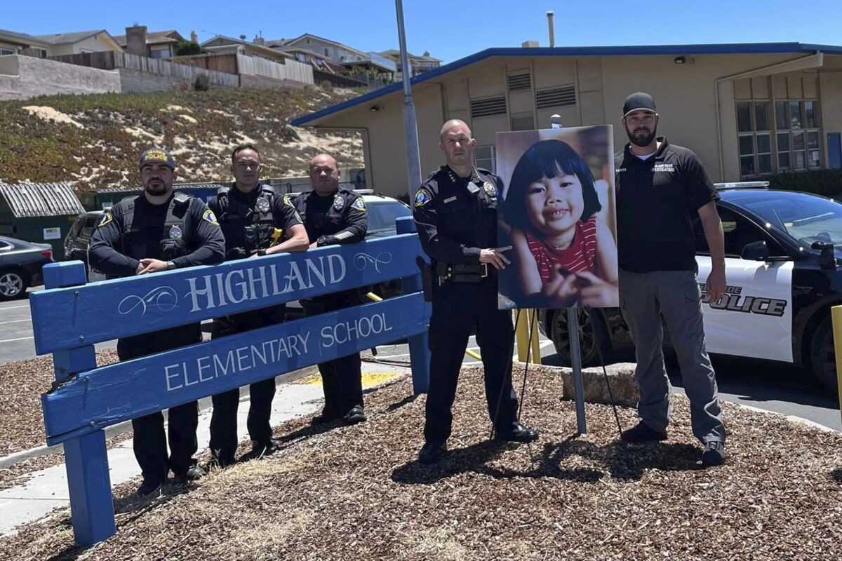 In this photo provided by the Seaside Police Department, interim Police Chief Nick Borges, second from right, and Detective Joshua Parker, right, stand next to a photo of Anne Pham outside Highland Elementary School in Seaside, Calif., on July 7, 2022. A 70-year-old Nevada man has been charged in the 1982 killing of the 5-year-old girl who disappeared while walking to her kindergarten class. Robert John Lanoue, of Reno, Nevada, was charged in the killing of Anne Pham and was due in court Monday, July 11, 2022, in Washoe County for a hearing about his extradition to Monterey County in California. (Seaside Police Department via AP)