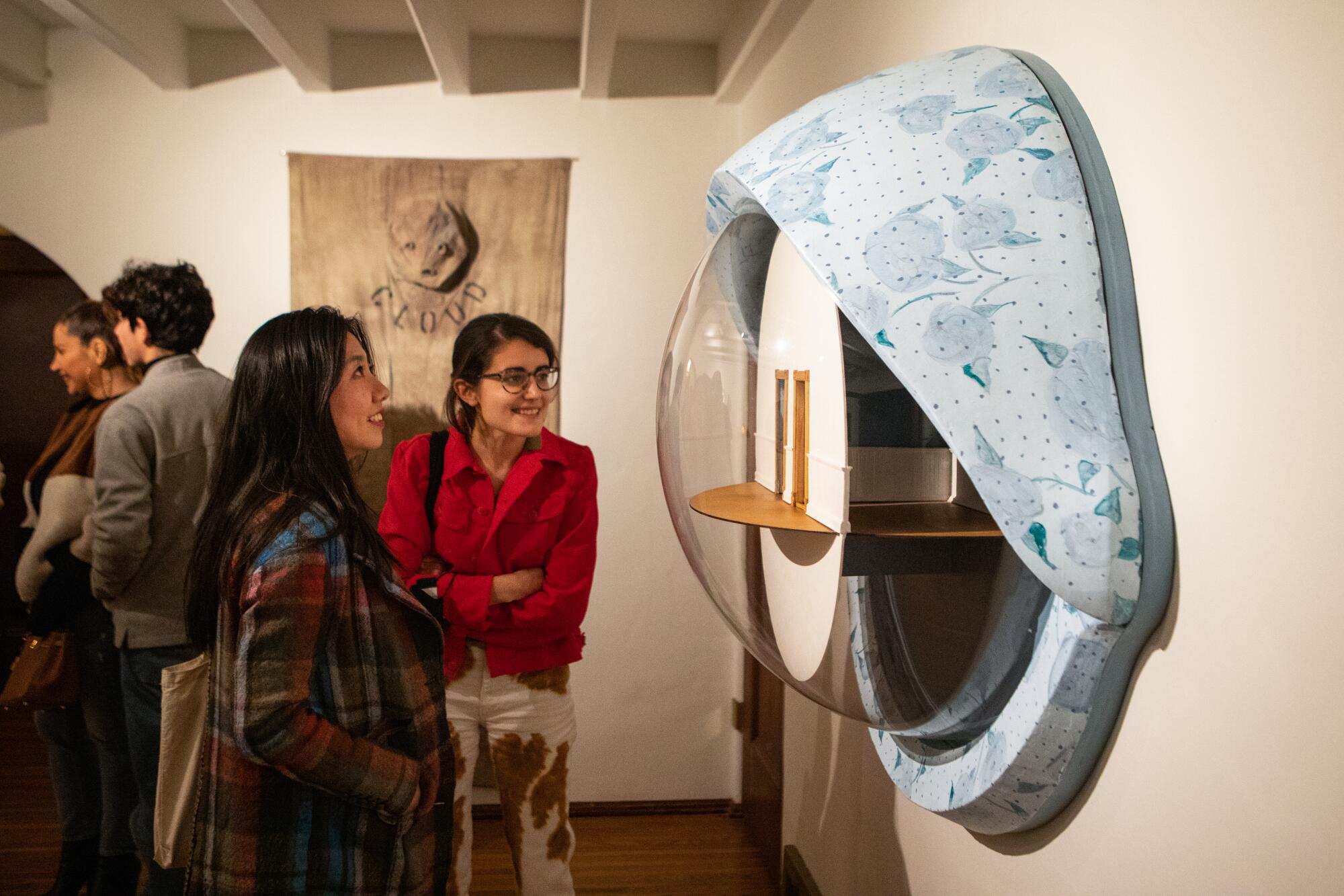 Two women look at an art piece hanging on a wall. It resembles a large, open eye.