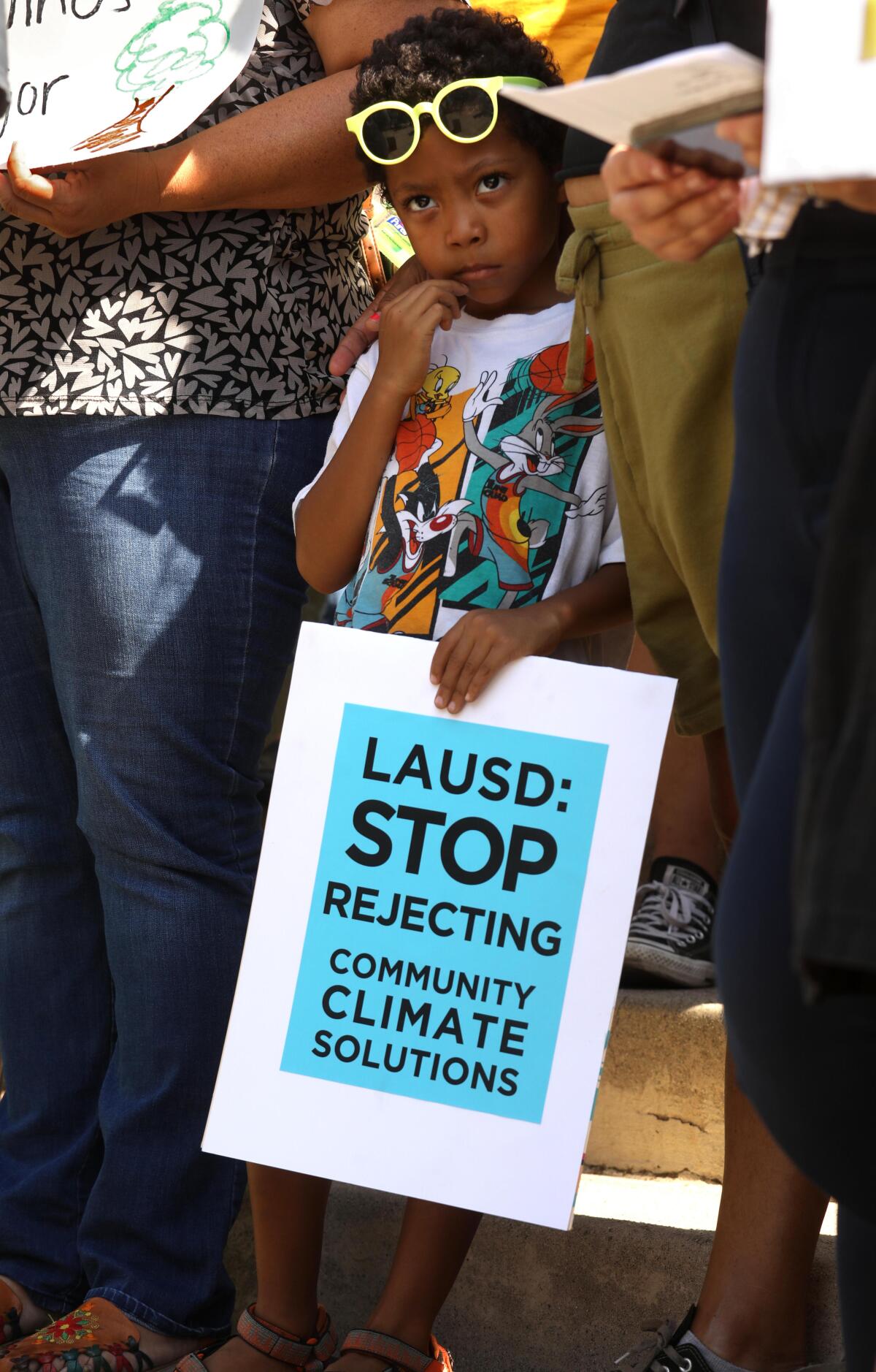 A child stands between two adults. The child is holding a sign that says, "LAUSD: Rejecting community climate solutions."