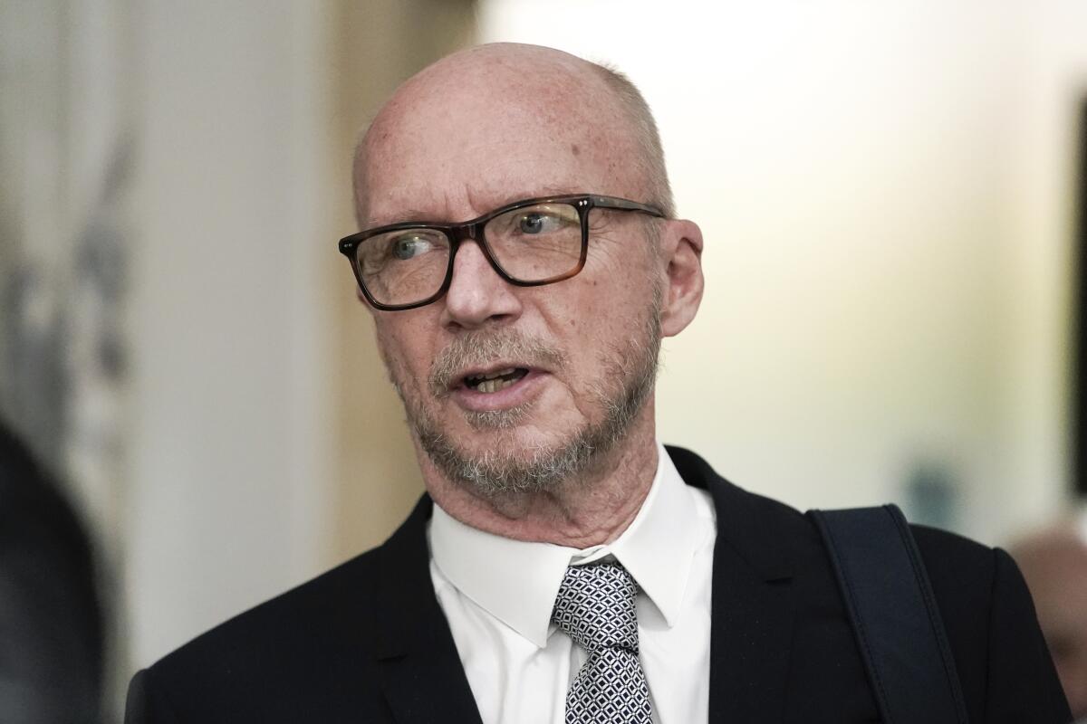 A balding man with glasses wearing a suit and tie talks to someone off camera