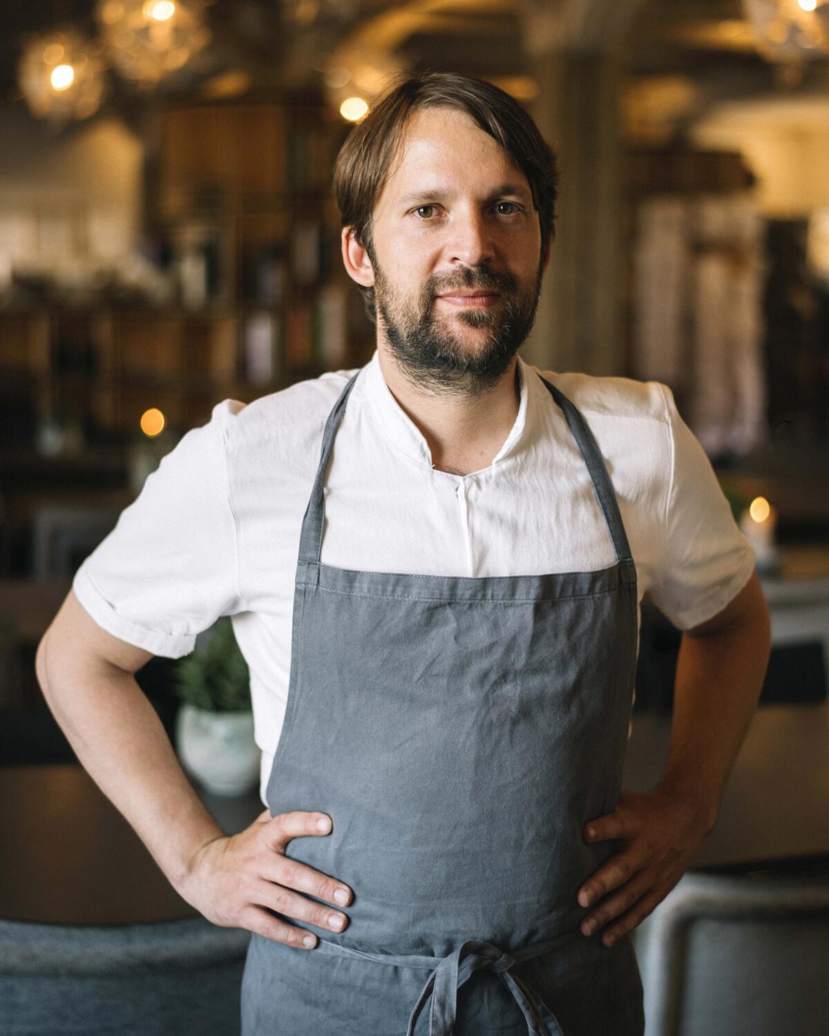 A bearded man in a white shirt and an apron, hands on hips 