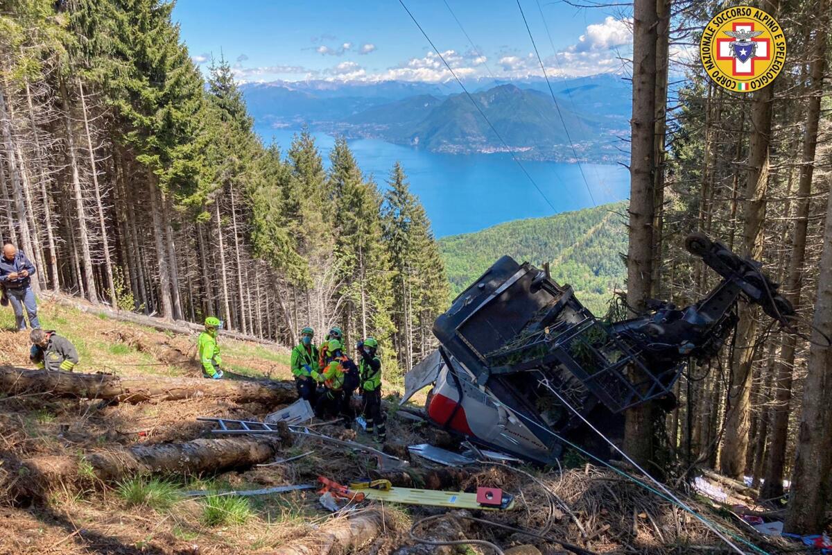 On a tree-covered mountainside, with a lake and mountains in the distance, rescuers stand alongside wreckage and gear.